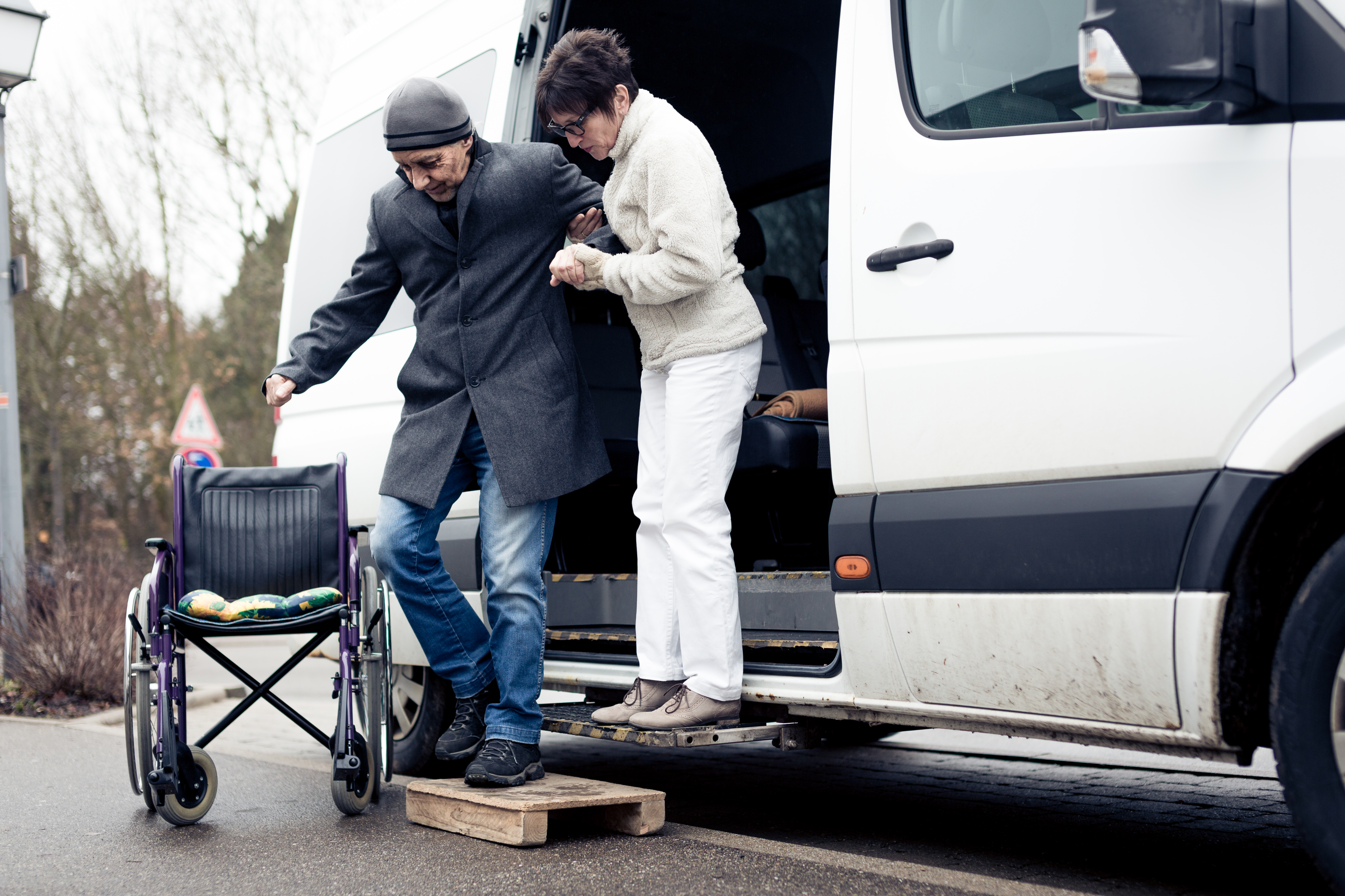 A man is exiting a transit vehicle with assistance from a woman through the slide door open on a transit vehicle, and he is using a step to make way towards a wheelchair on the ground.