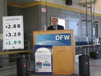 This is a photo of a man at a DFW labeled podium with a sign displaying decreasing gas prices.